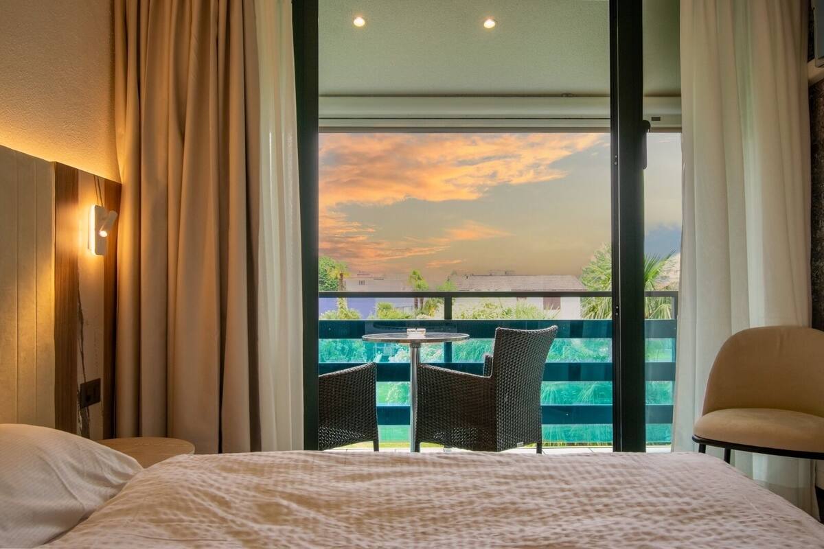 A hotel room with a bed, a small table, two chairs, and a balcony with a view of the sunset.