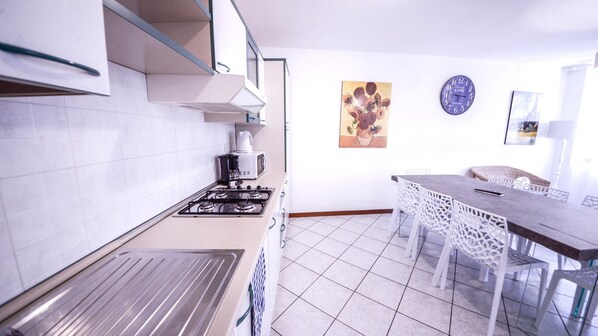 A modern kitchen with white tiles, a gas stove, and a microwave. There is a dining area with a table and chairs.