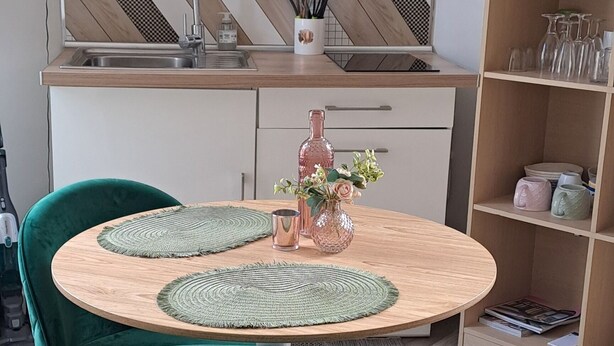 Et moderne køkken med et hvidt skab, en træbordplade og en indbygget ovn. Der er et rundt spisebord med grønne polstrede stole og en vase med blomster.