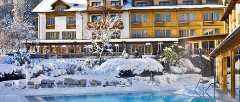 A hotel with a pool surrounded by snow, featuring a large building with multiple balconies and a mountain backdrop.