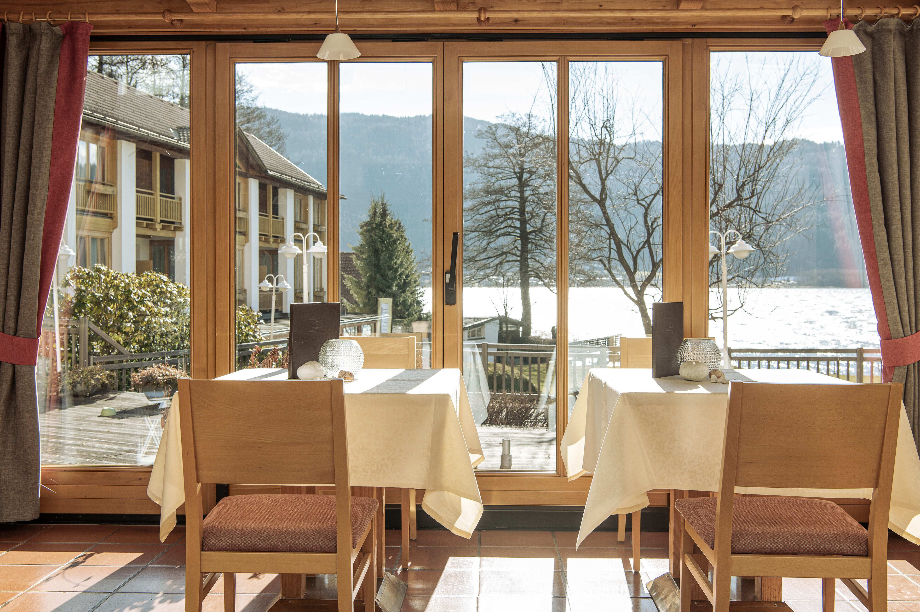 Un espace repas comprenant deux tables et des chaises, de grandes fenêtres offrant une vue sur un lac et des montagnes, et un plafond en bois.