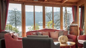 Un salon chaleureux avec un canapé rouge, des fauteuils gris et une table basse en bois. De grandes fenêtres offrent une vue sur un lac et des montagnes.