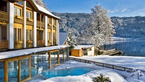Un hôtel doté d’une piscine à ciel ouvert protégée par des parois de verre, de balcons en bois et offrant une vue sur un paysage enneigé et un lac.