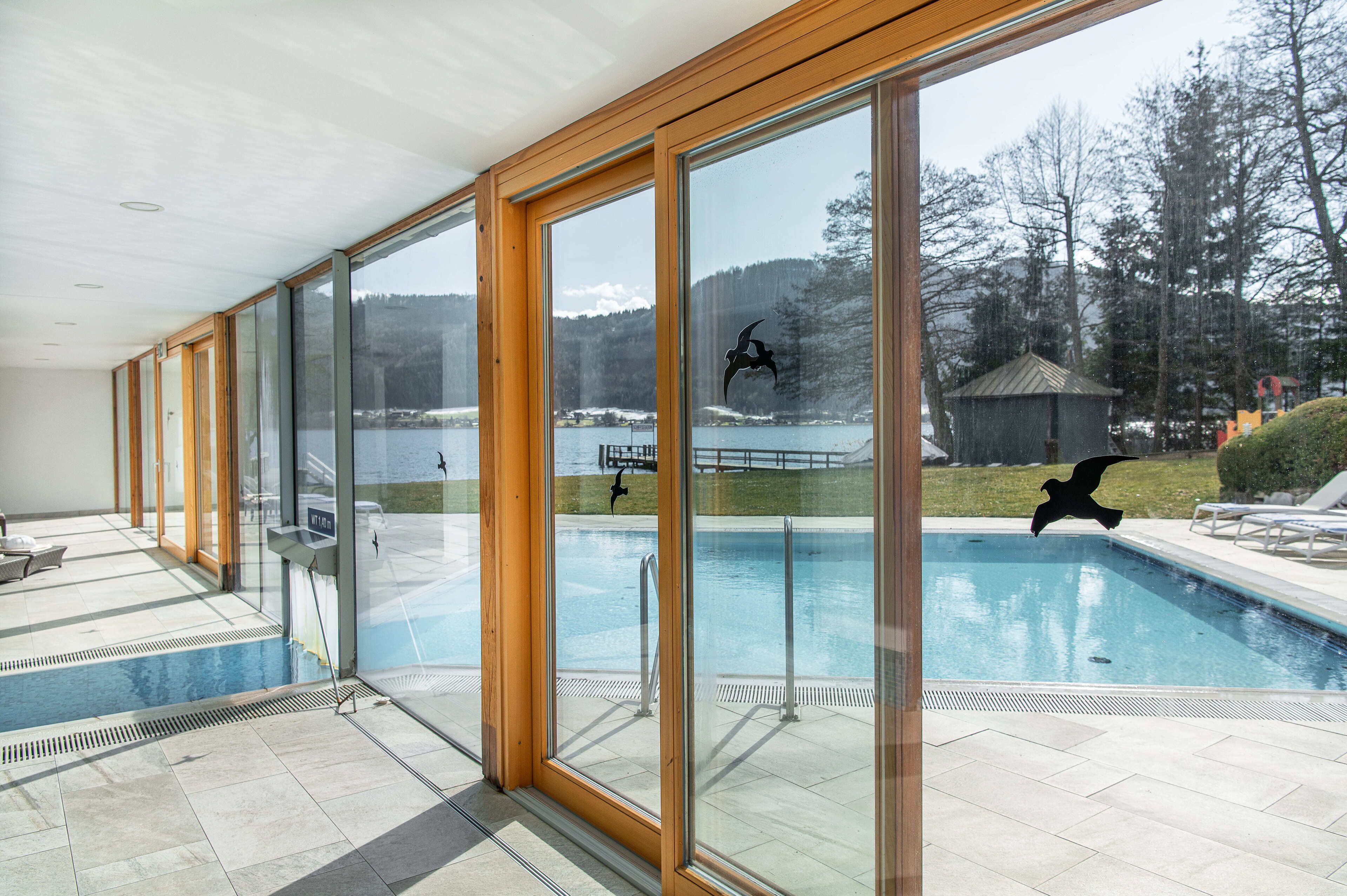 A swimming pool with clear glass walls, a view of a lake, and a small building in the background.