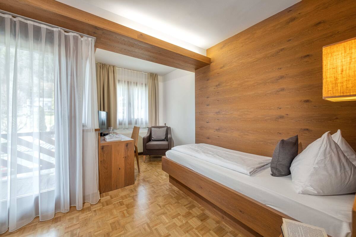 Een moderne kamer met houten vloer, een bed met kussens, een bureau met stoel en dunne gordijnen.