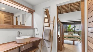 Une salle de bain avec un meuble-lavabo en bois, un grand miroir et une vue sur une chambre à coucher avec un lit et une fenêtre.