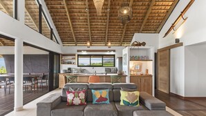 Un intérieur moderne avec un plafond en bois, un canapé gris agrémenté de coussins colorés, un coin cuisine et une vue dégagée sur l’extérieur grâce à une grande fenêtre.
