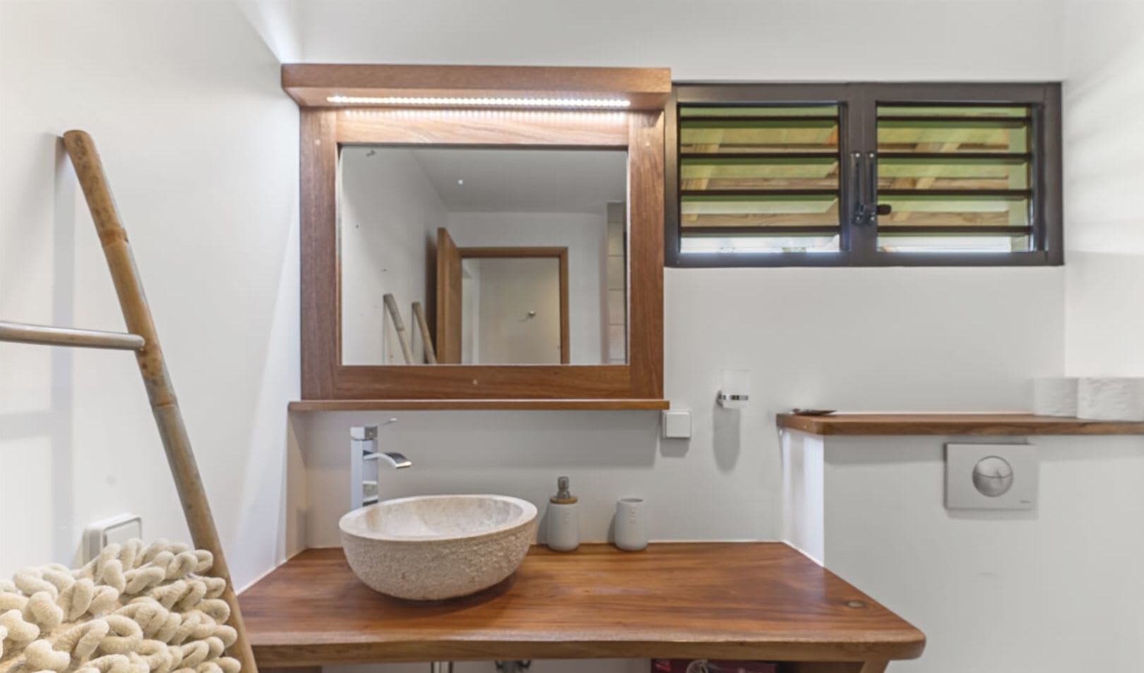 Un baño con tocador de madera, lavabo de piedra, espejo, ventana con persianas y un perchero para toallas.