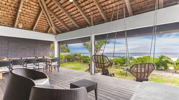 A covered outdoor area with wicker furniture, a table, and chairs, overlooking a beach.