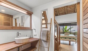 Un baño con tocador de madera, un espejo grande y vista a un dormitorio con cama y ventana.