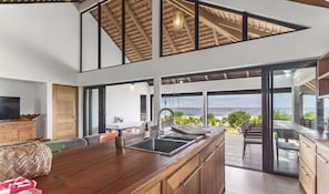 Een moderne keuken met een groot eiland, een eetgedeelte met uitzicht op de oceaan, en een ruime woonkamer met hoge plafonds en grote ramen.
