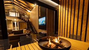Un interior moderno con una mesa de comedor de madera, un cuenco negro grande con fuego, un sofá y un televisor montado en una pared de madera.