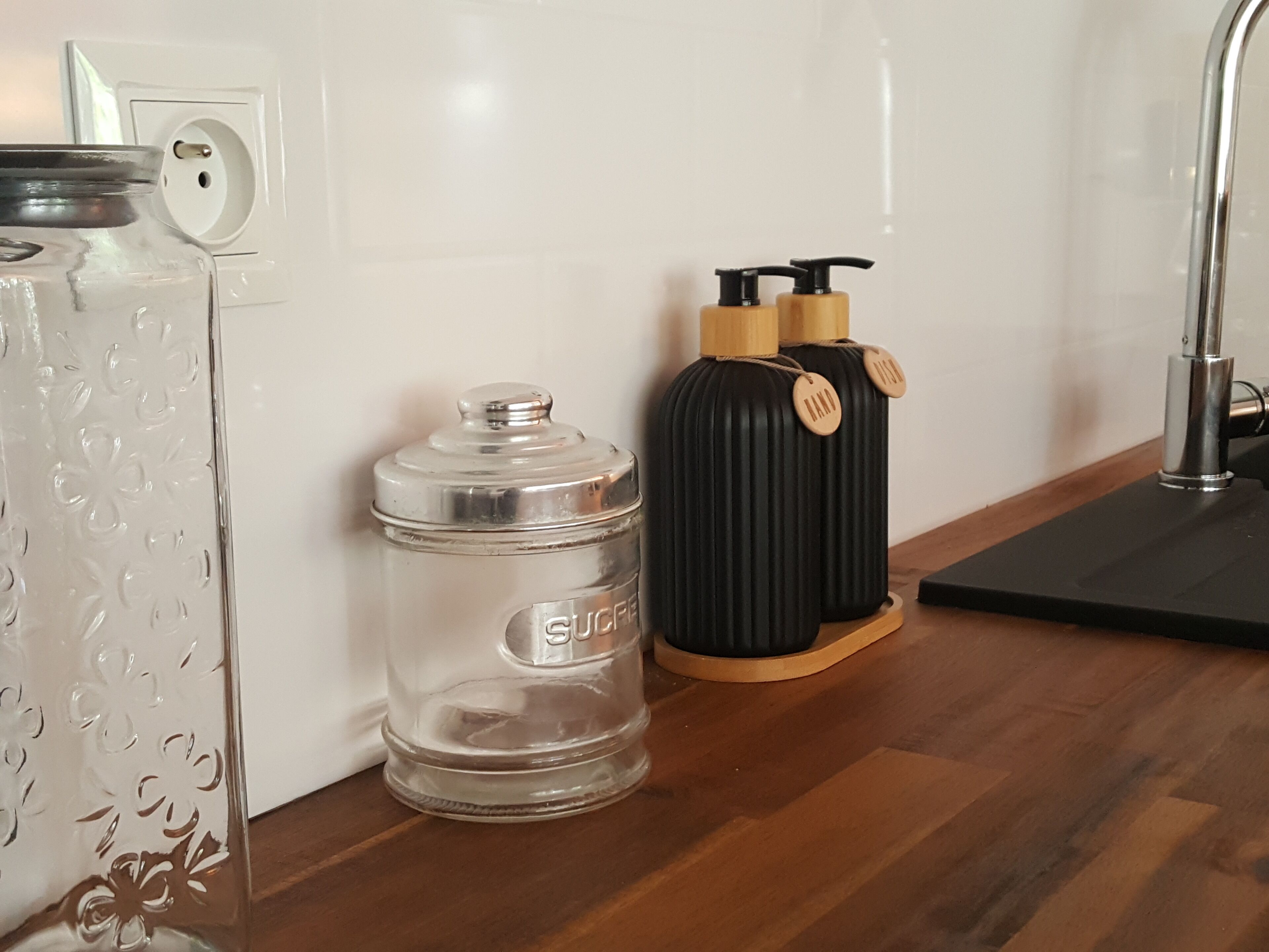 Una encimera de cocina con un recipiente de vidrio etiquetado “SUCRE”, un recipiente negro con asas de madera y un grifo metálico.
