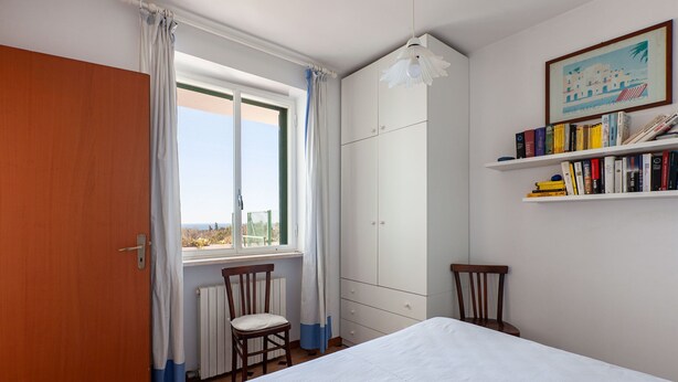 A bedroom with a bed, a wooden door, a window with curtains, a radiator, a chair, a bookshelf, and a framed picture on the wall. - Villa Giorgisa Apartment 2 Pool Sea View (Ugento)