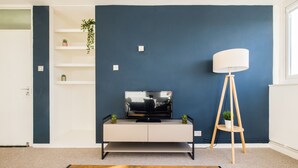 Een moderne woonkamer met een donkerblauwe accentmuur, een witte tv-meubel met een flatscreen-televisie, een houten vloerlamp en een klein plantje op een plank.