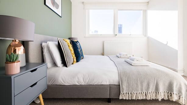 Een moderne slaapkamer met een bed, nachtkastje, lamp en een raam met uitzicht op gebouwen.