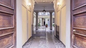 Un pasillo con puertas de madera y columnas que conduce a un patio con plantas en macetas y vistas a edificios.