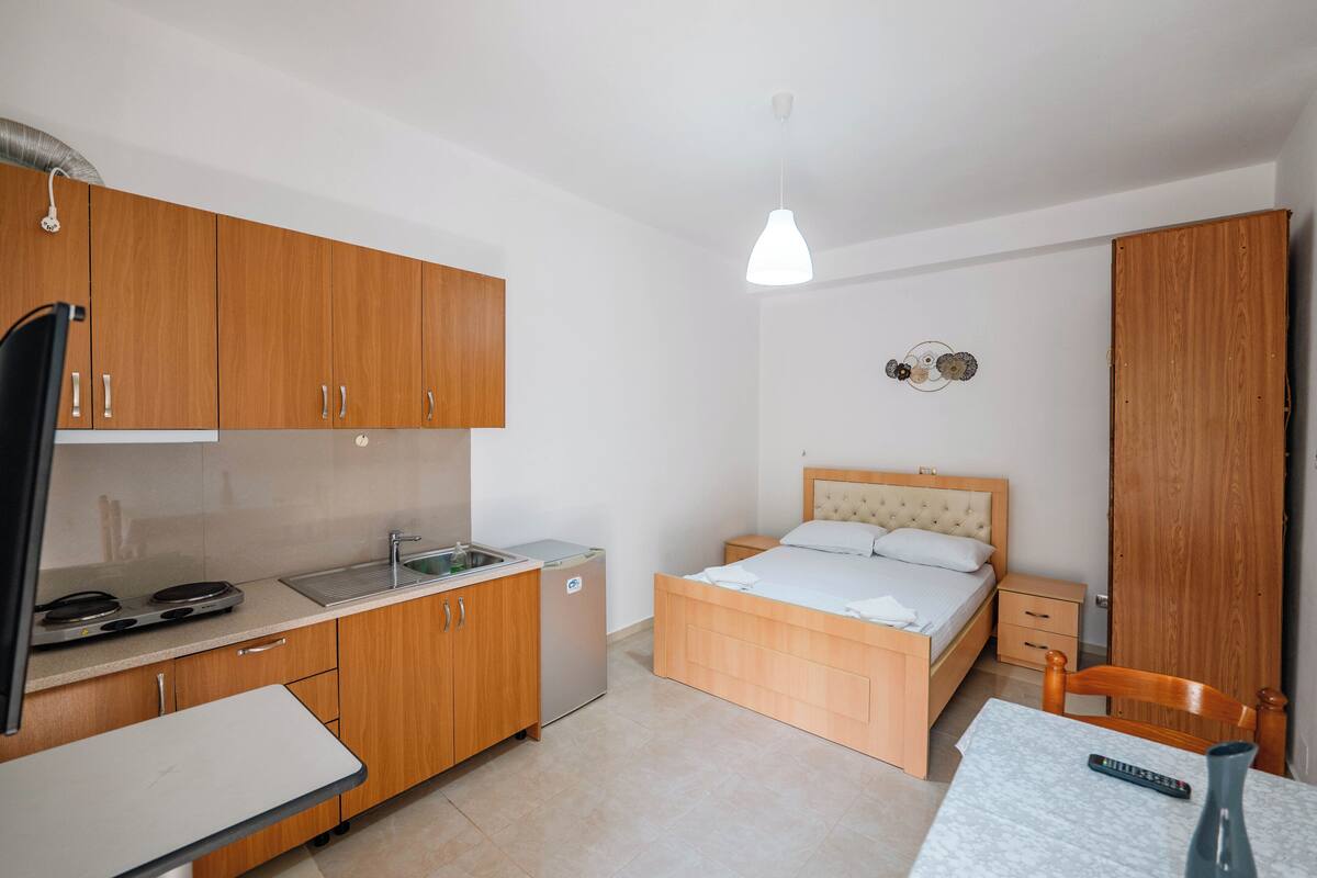 Una habitación compacta con kitchenette, una cama con cabecera de madera, una mesita con un control remoto y un adorno en la pared.