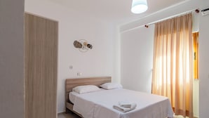 Una cama bien hecha con sábanas blancas y una toalla doblada, un cabecero de madera, una lámpara de pared y una ventana con cortinas traslúcidas.