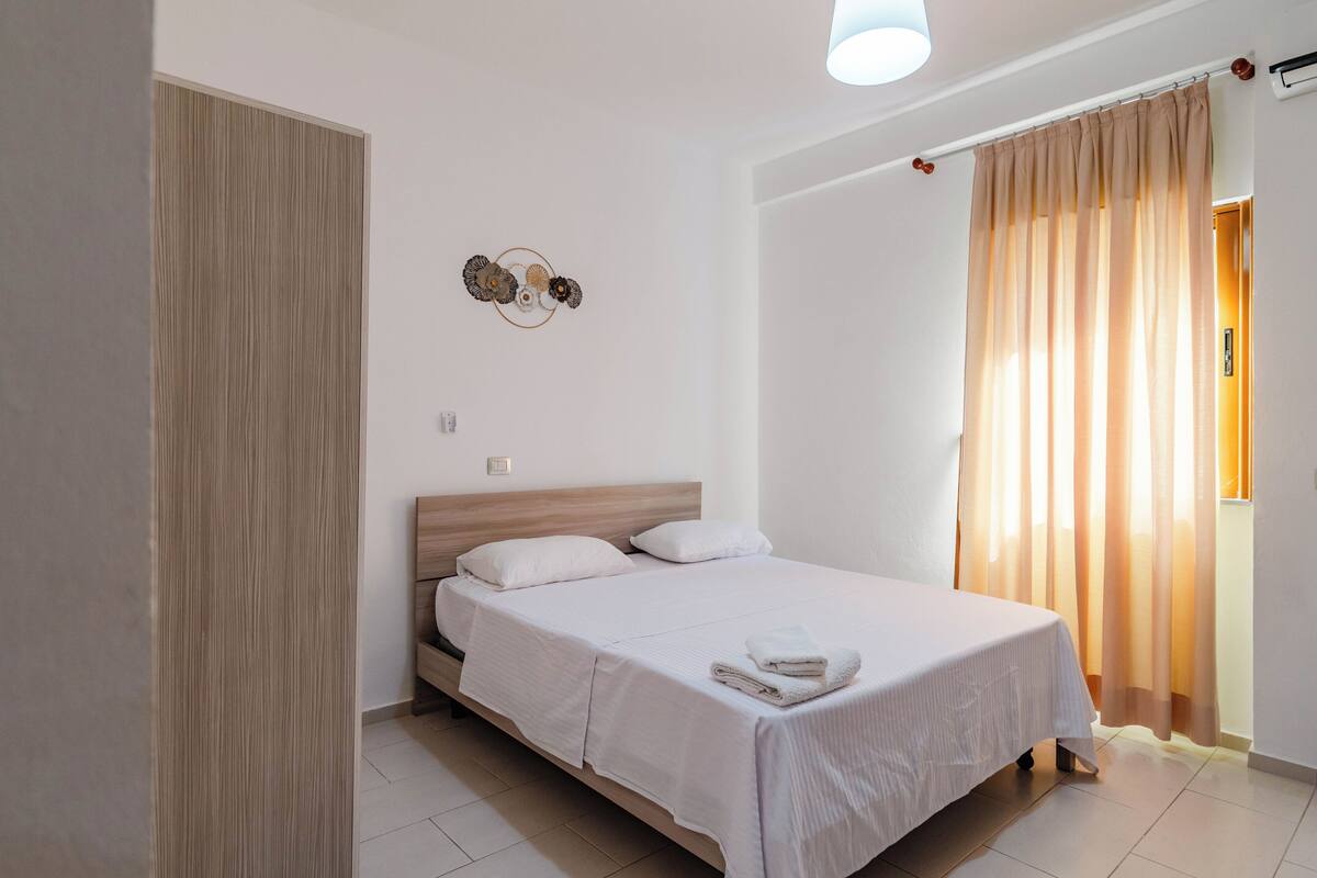 Una cama bien hecha con sábanas blancas y una toalla doblada, un cabecero de madera, una lámpara de pared y una ventana con cortinas traslúcidas.