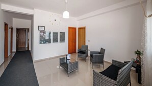 Una sala de estar moderna con una mesa de centro de vidrio, sillas de mimbre, una lámpara colgante blanca y una vista urbana enmarcada en la pared.