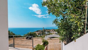 Vista desde un balcón sobre una zona costera con cielo despejado, una moto estacionada al frente y un muro blanco con valla de madera y follaje.