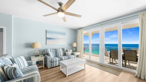 A room with a large window, a ceiling fan, a striped sofa, a coffee table, and a view of the ocean. - Luxurious 4BD! Oceanview! Free Beach Chair Service! (Panama City Beach)