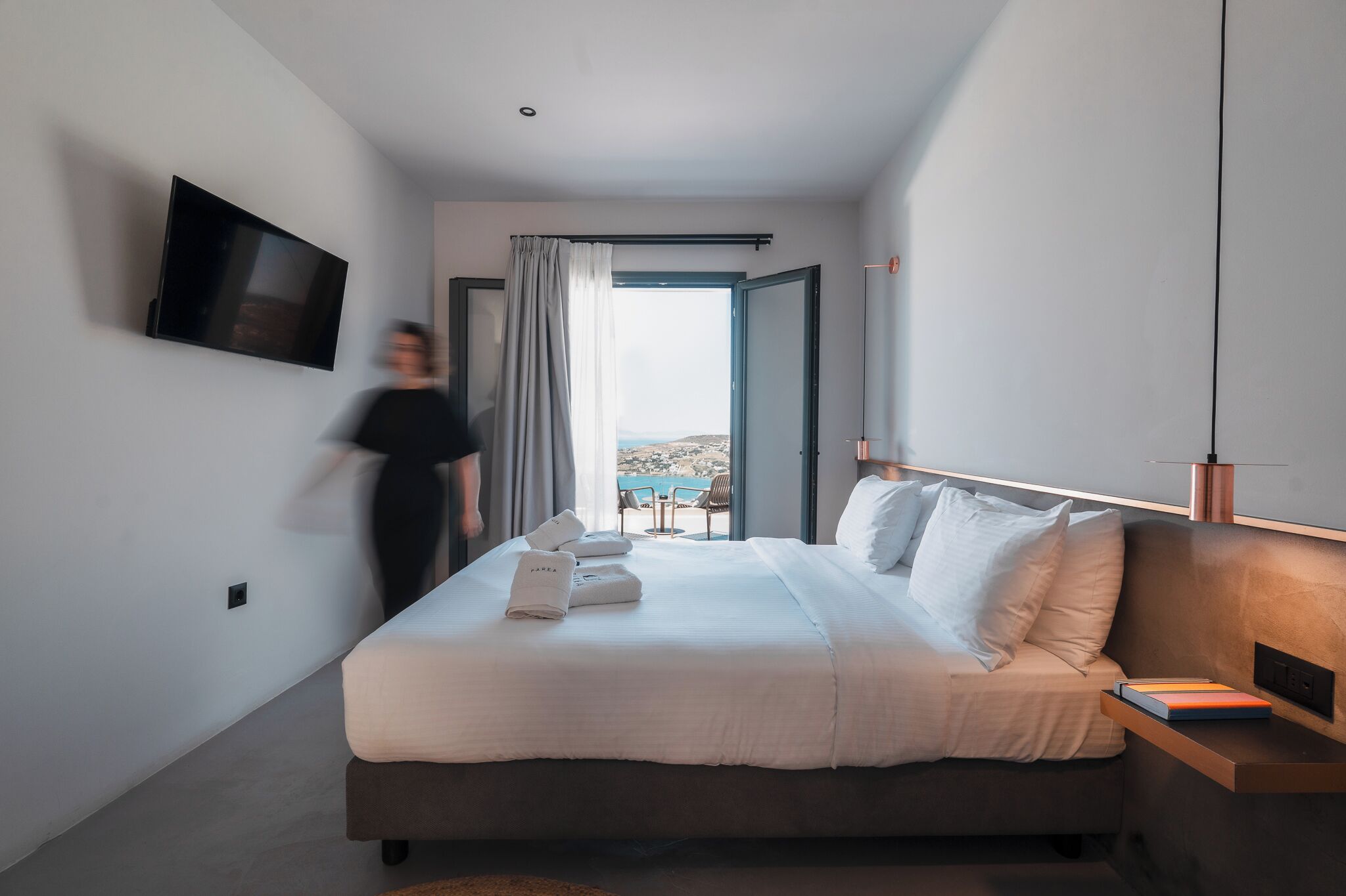 A modern hotel room with a large bed, a flat-screen TV mounted on the wall, and a view of the outdoors through a large window.