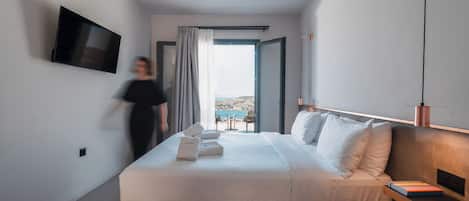 A modern hotel room with a large bed, a flat-screen TV mounted on the wall, and a view of the outdoors through a large window.