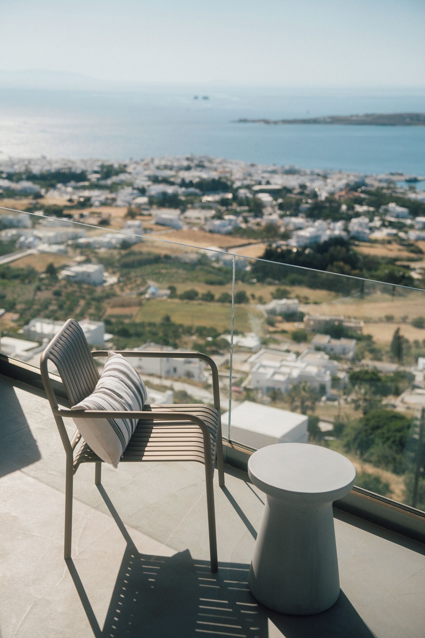 A balcony with a chair and a small table, overlooking a coastal town with buildings and greenery.
