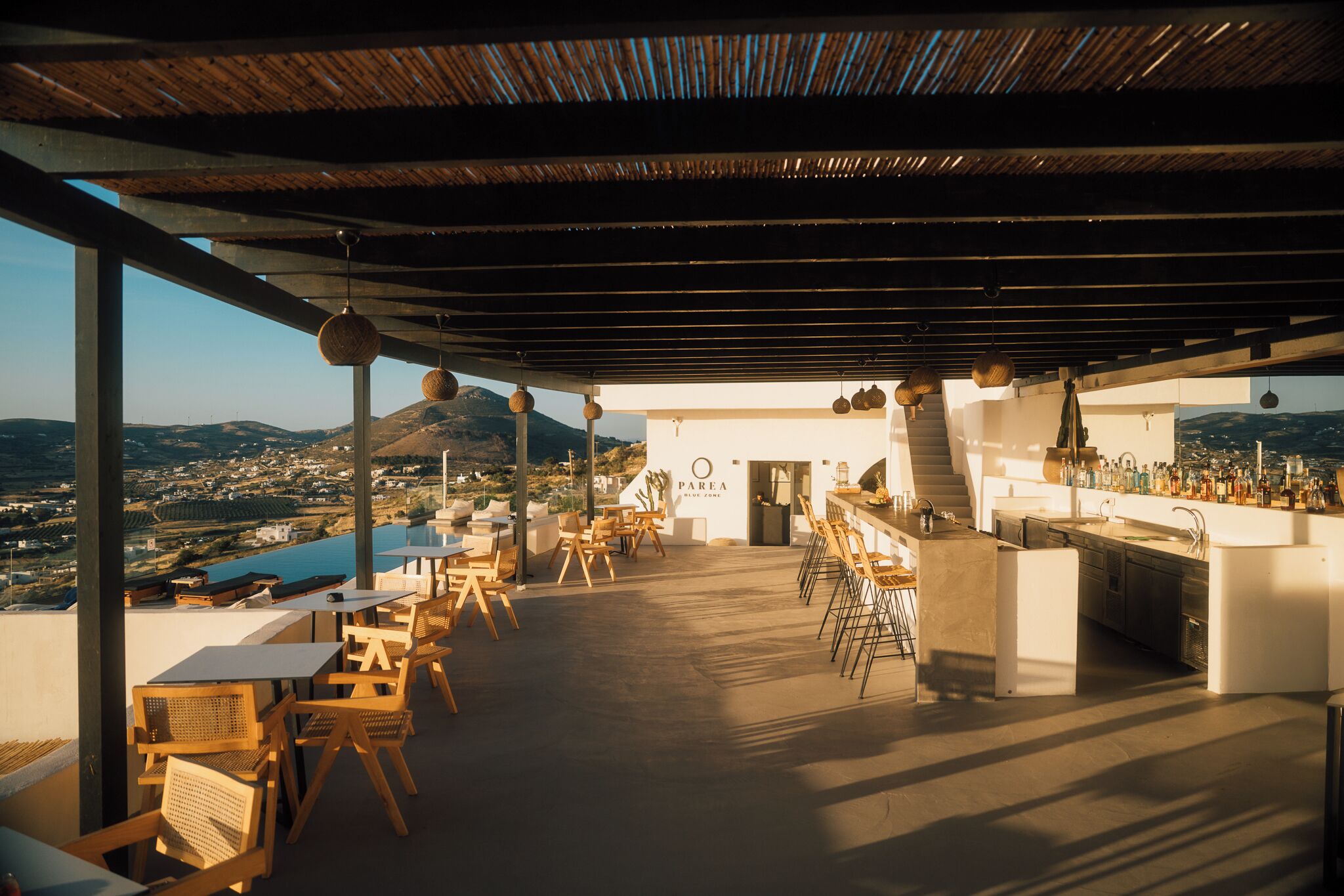 Un bar en la azotea con mesas y sillas de madera, una barra y vistas a la ciudad y las colinas.