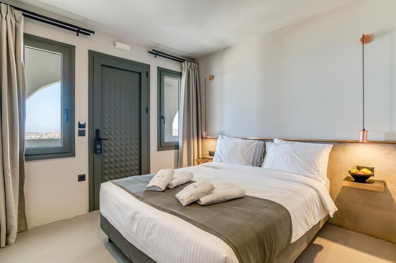A hotel room with a large bed, two doors, a window with curtains, a bedside table with a lamp, and a view of the city.