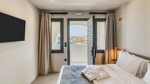 Ένα δωμάτιο ξενοδοχείου με ένα κρεβάτι, μια τηλεόραση τοποθετημένη στον τοίχο και θέα στη θάλασσα από μια ανοιχτή πόρτα.