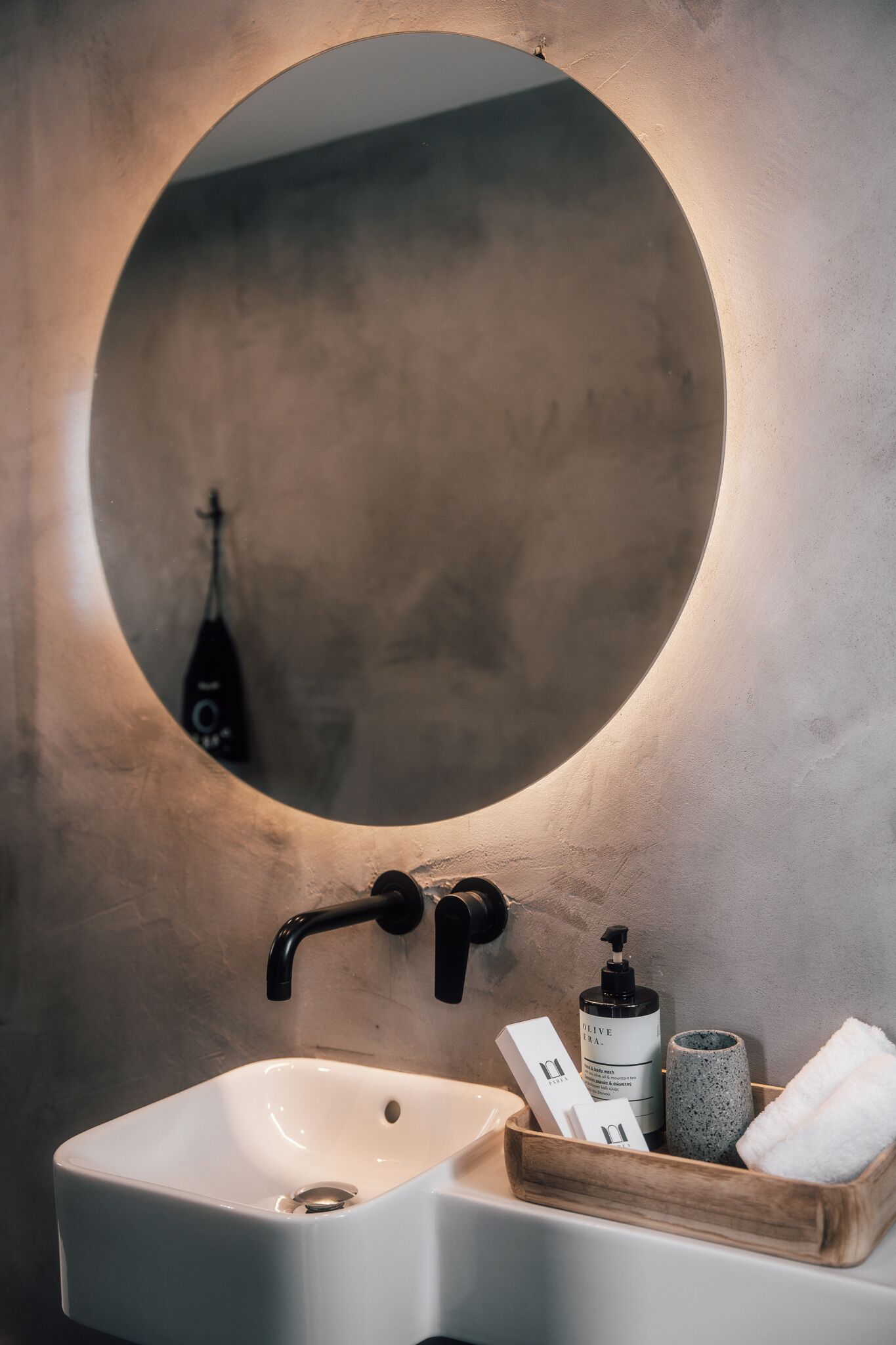 A bathroom with a round mirror, a white sink, a wooden tray with toiletries, and a bottle of olive oil.