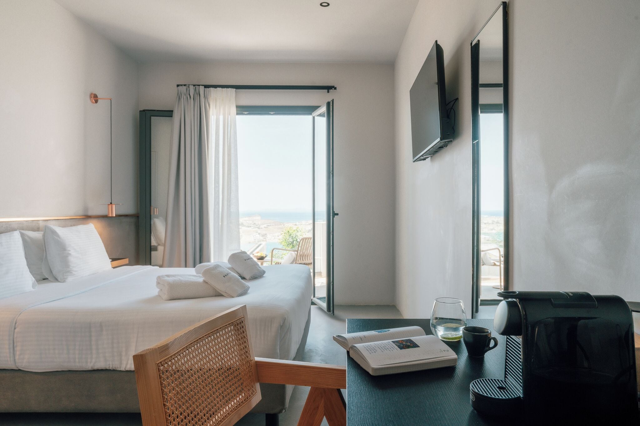 Quarto de hotel com cama, escrivaninha com cafeteira, um livro e vista para o exterior.