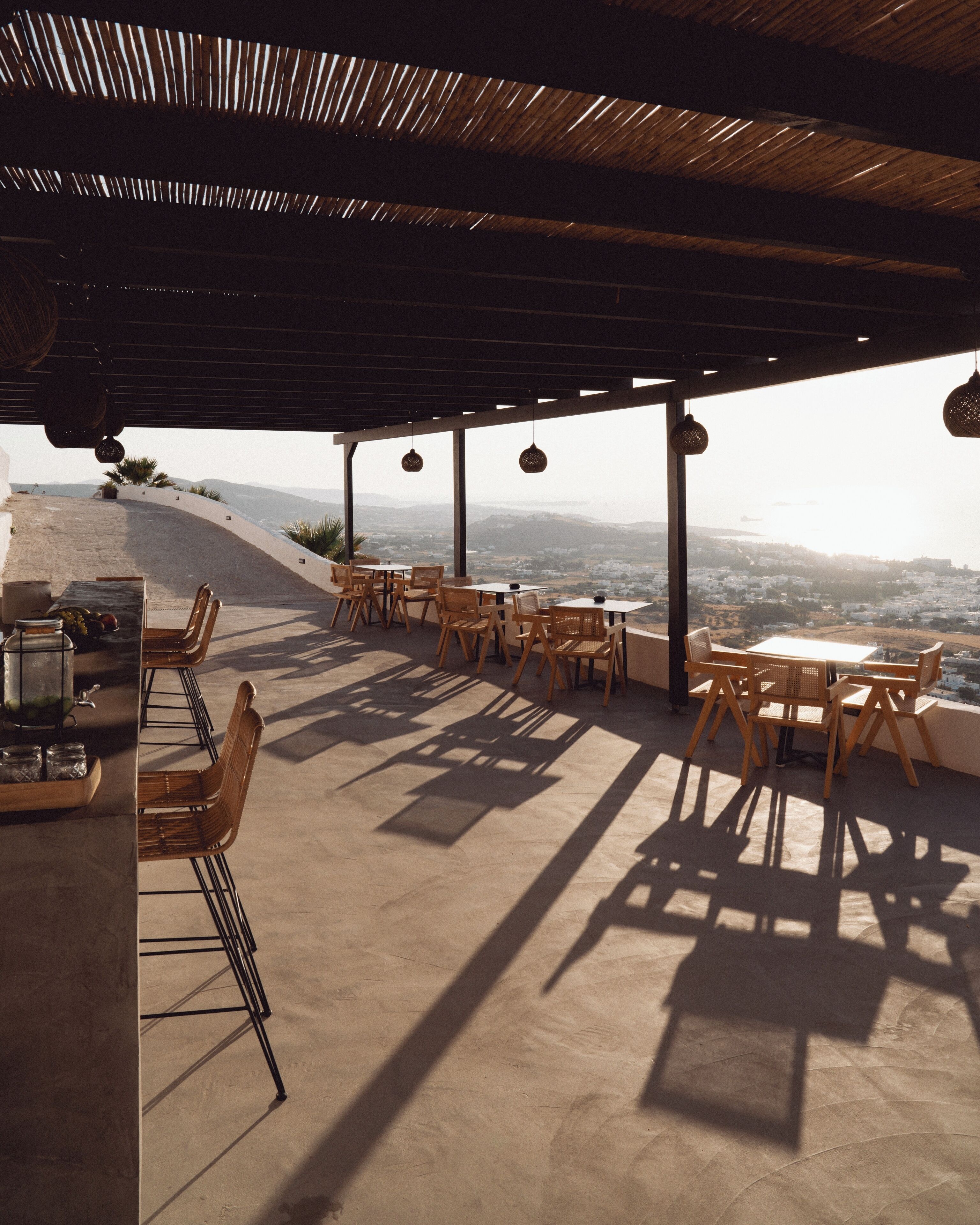 An outdoor patio with wooden furniture and hanging lights, overlooking a cityscape.