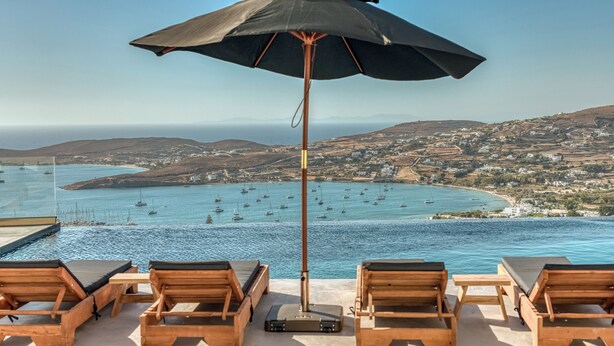 Ένας χώρος δίπλα στην πισίνα με ξύλινες ξαπλώστρες και μια μεγάλη ομπρέλα, με θέα σε μια παραθαλάσσια πόλη και κρυστάλλινα, γαλάζια νερά.