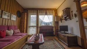 Ein gemütliches Wohnzimmer mit Holzwänden, einer Couch, einem Couchtisch mit Schalen, einem Fernseher und Blick auf Berge durch das Fenster.