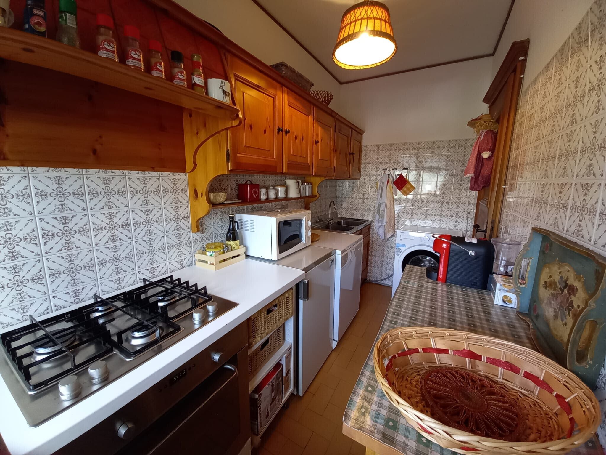 Een compacte keuken met een gasfornuis, magnetron en wasmachine. Er zijn houten keukenkastjes en een betegelde spatwand.