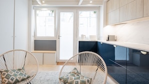 Une cuisine moderne avec des meubles blancs, deux chaises en osier ornées de coussins à motifs et une vue à travers une porte vitrée.