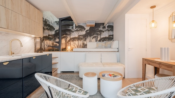 Une cuisine moderne avec un comptoir en marbre, un robinet doré et une fresque représentant des arbres sur le mur. On y trouve deux chaises en osier blanches, une petite table ronde et une table d’appoint en bois.