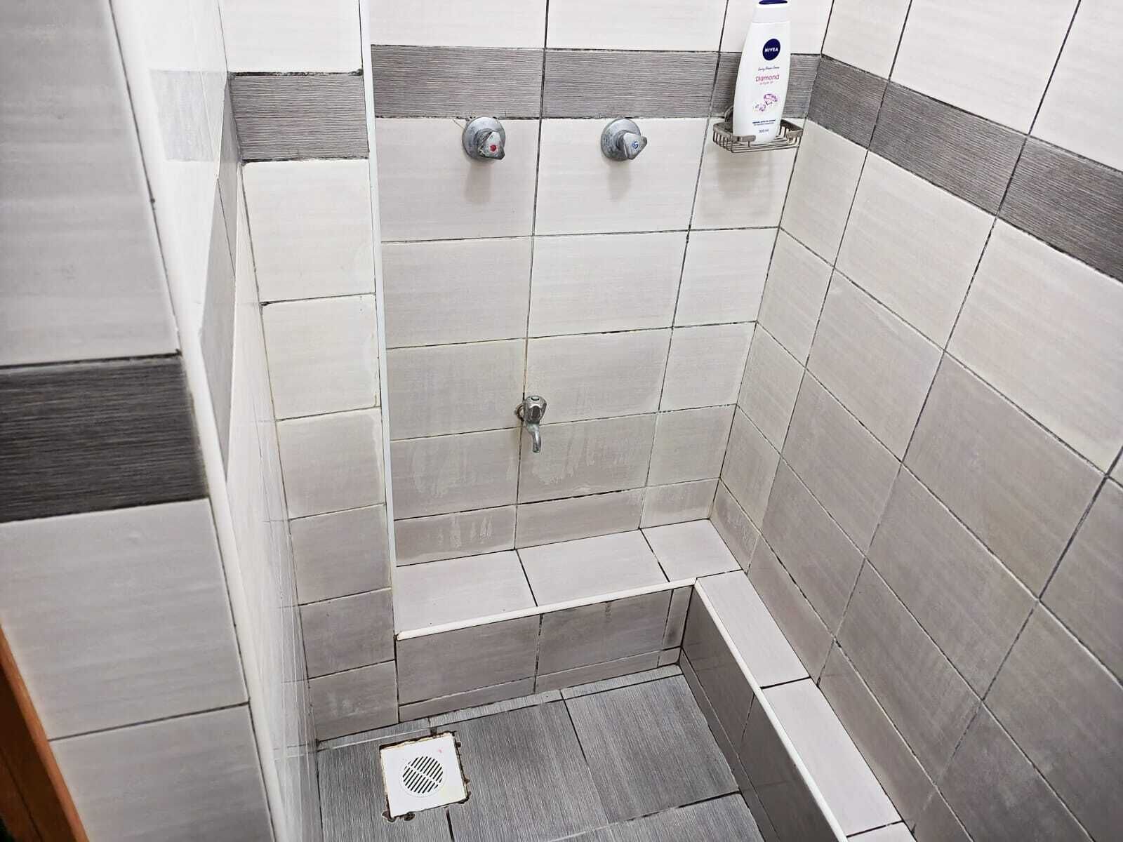 Un área de ducha con azulejos grises y blancos, un cabezal de ducha manual y una botella de champú en la pared.