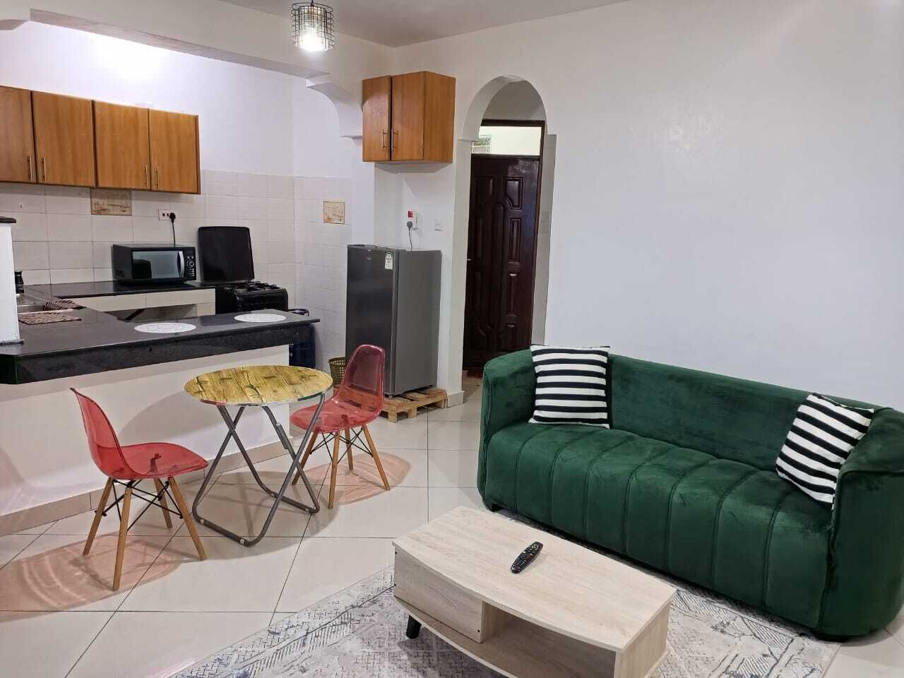 A compact living space with a green sofa, a small dining table with chairs, a kitchen area with cabinets and appliances, and a door leading to another room.
