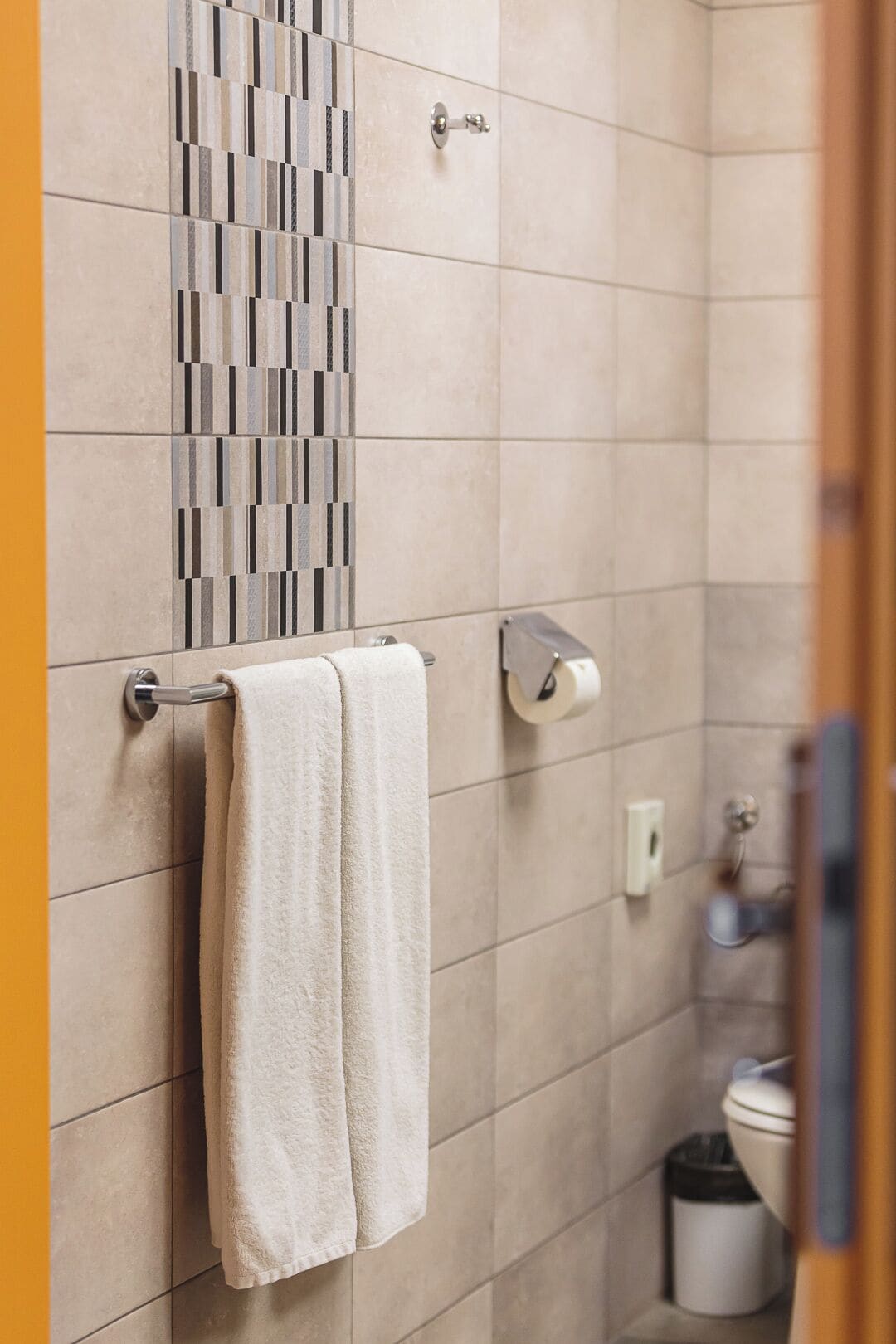 Un baño con baldosas color crema, un perchero con dos toallas, un dispensador de papel higiénico y un cubo de basura.