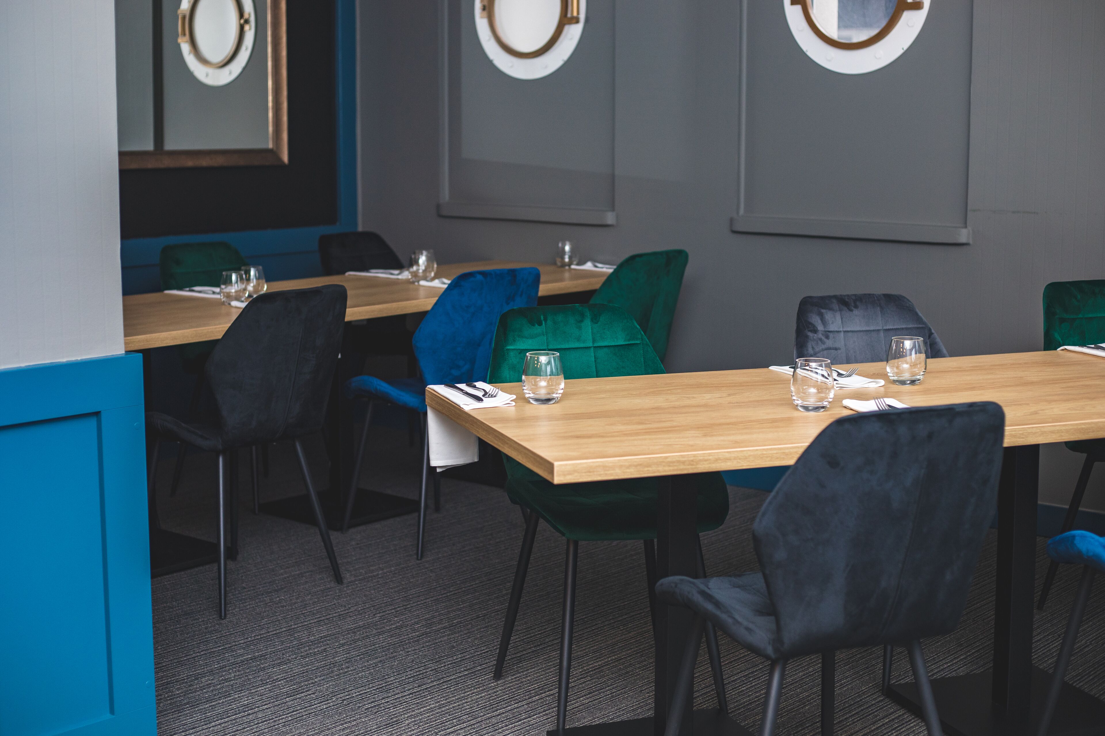 Un espace repas moderne avec des tables et des chaises en bois, dont certaines sont recouvertes d’un tissu vert et bleu.