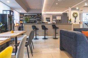 Un espace bar moderne avec un comptoir en bois, des tabourets de bar noirs et un coin salon comprenant un canapé bleu et un fauteuil jaune.