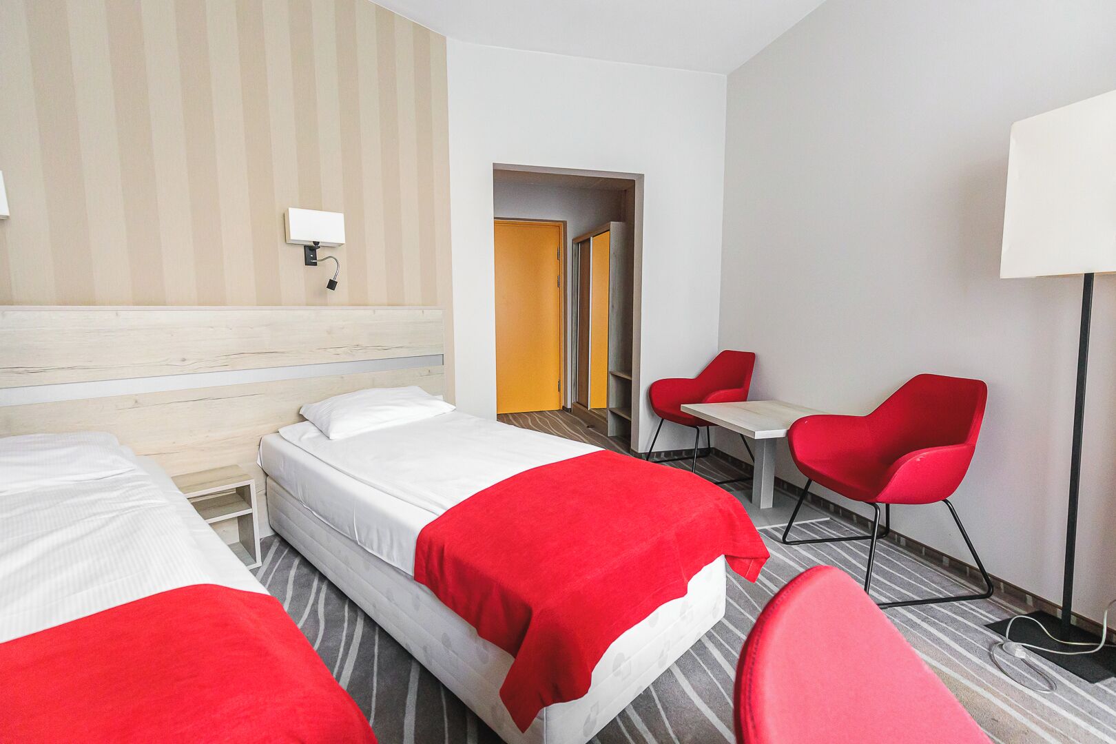 A hotel room with two beds, a desk, two red chairs, a lamp, and a view of a corridor.