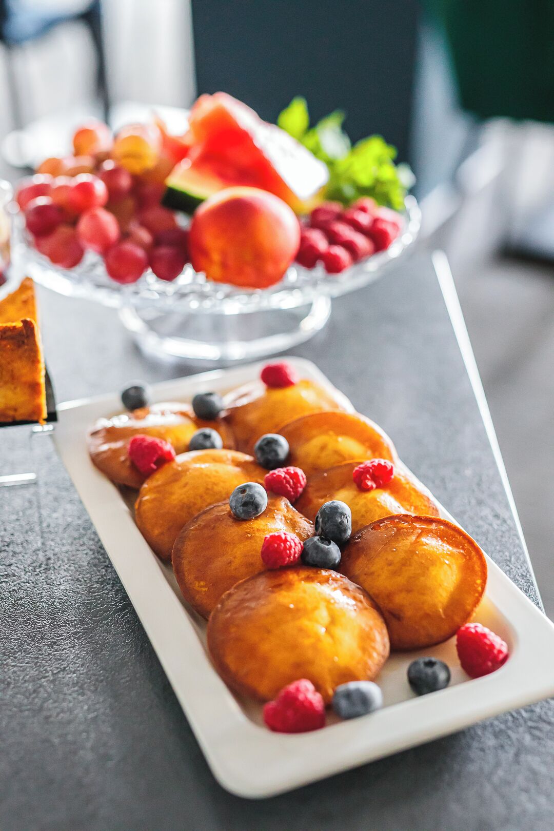 A plate of glazed donuts with blueberries and raspberries, and a bowl of assorted fruits in the background.