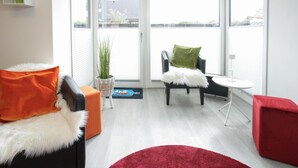 A modern living room with a white sofa, a red ottoman, a black chair with a green pillow, a small white table, and a red rug.