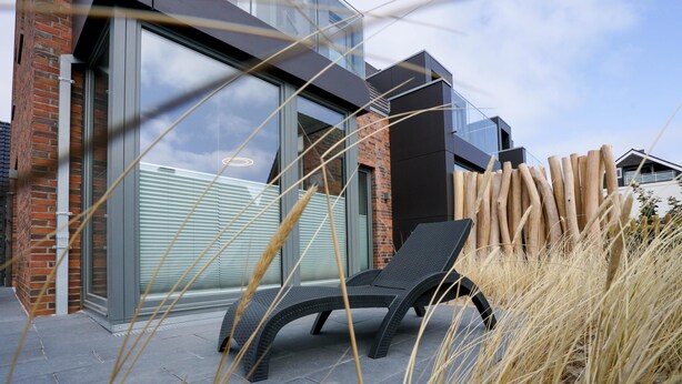 A modern building with large glass windows, a brick facade, and a lounge chair in the foreground.